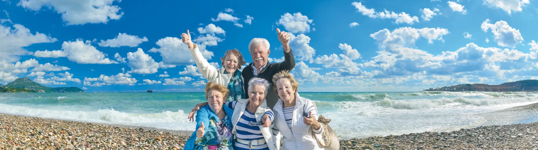 Lebensfreunde Dresden - Dieses Bild zeigt eine Gruppe von fünf Personen, die fröhlich auf einem Kieselstrand vor einem klaren blauen Himmel posieren.